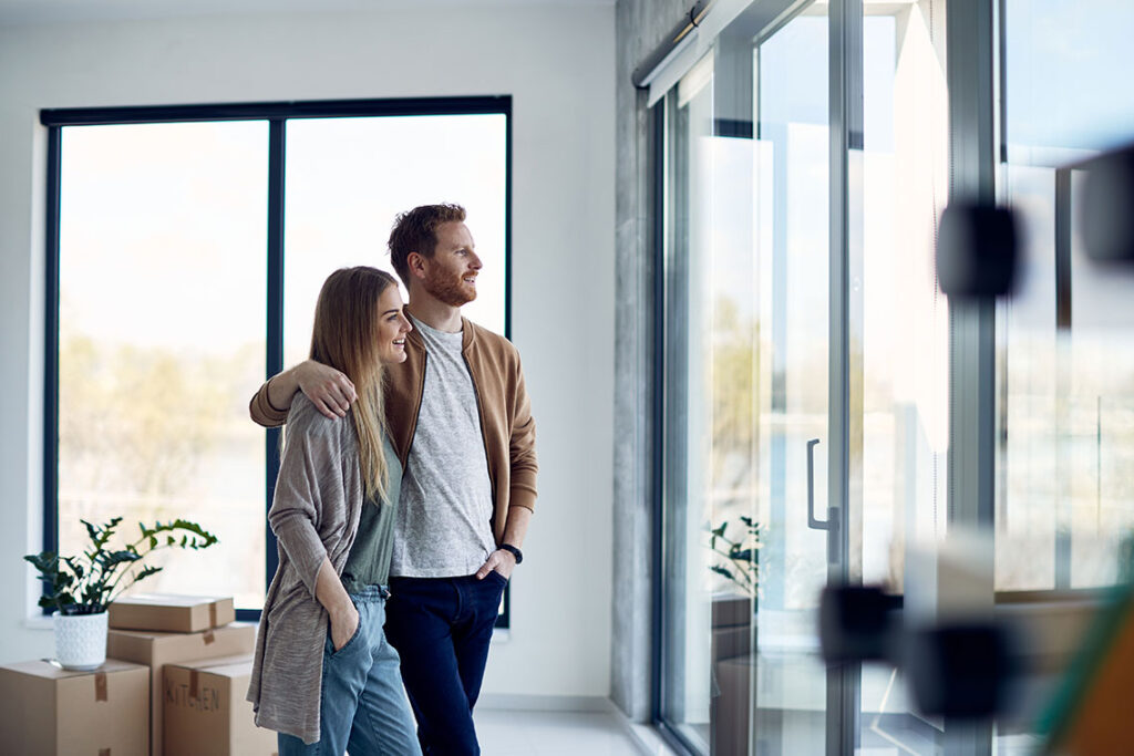 Ein glückliches junges Paar steht im Wohnzimmer ihrer eigenen Immobilie und sieht aus dem Fenster.